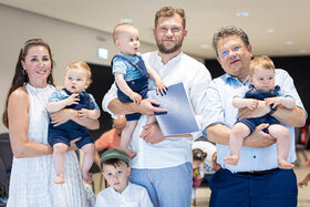 Eine Mutter, ein Vater und ein Mann halten drei kleine Kinder auf den Armen. Ein Junge steht vor der Gruppe. 